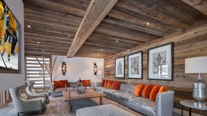 En rummelig stue med træloft, en grå sofa med orange puder, et marmorbord og et stort billede på væggen.