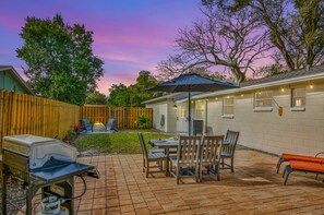 Une terrasse à l’arrière de la maison, équipée d’un barbecue, d’une table, de chaises et d’un parasol.