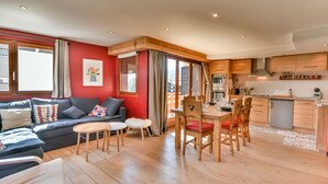 A modern living room with a sofa, a dining table, and chairs. There is a kitchen area with wooden cabinets and a refrigerator. - Stunning Renovated Mountain-Style 2BR+Cabin Apt near Village Center, Sleeps 6 (Les Gets)