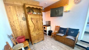 Een gezellige woonkamer met een houten kast, een bank met kussens met een hartmotief, een klein tafeltje en een aan de muur bevestigde televisie.