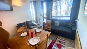 Een gezellige woonkamer met een houten eettafel gedekt voor twee personen, een donkergrijze bank en uitzicht op bomen door grote ramen.