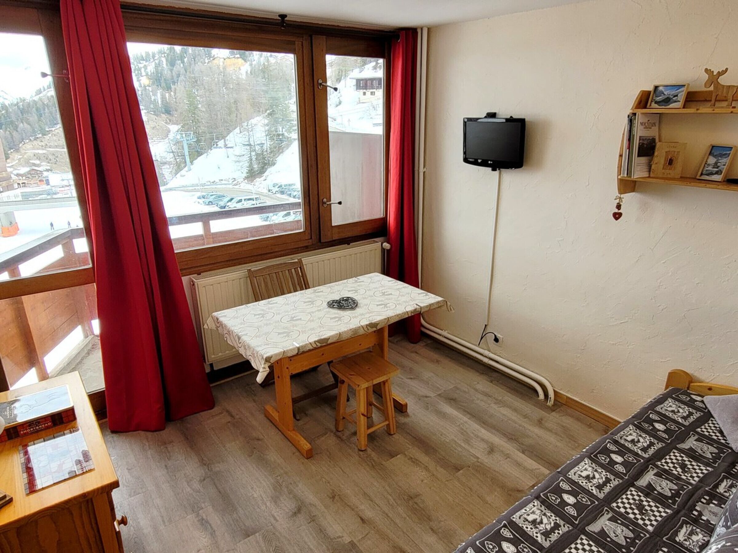 A room with a wooden table and chairs, a TV mounted on the wall, and a window with a view of a snowy landscape.