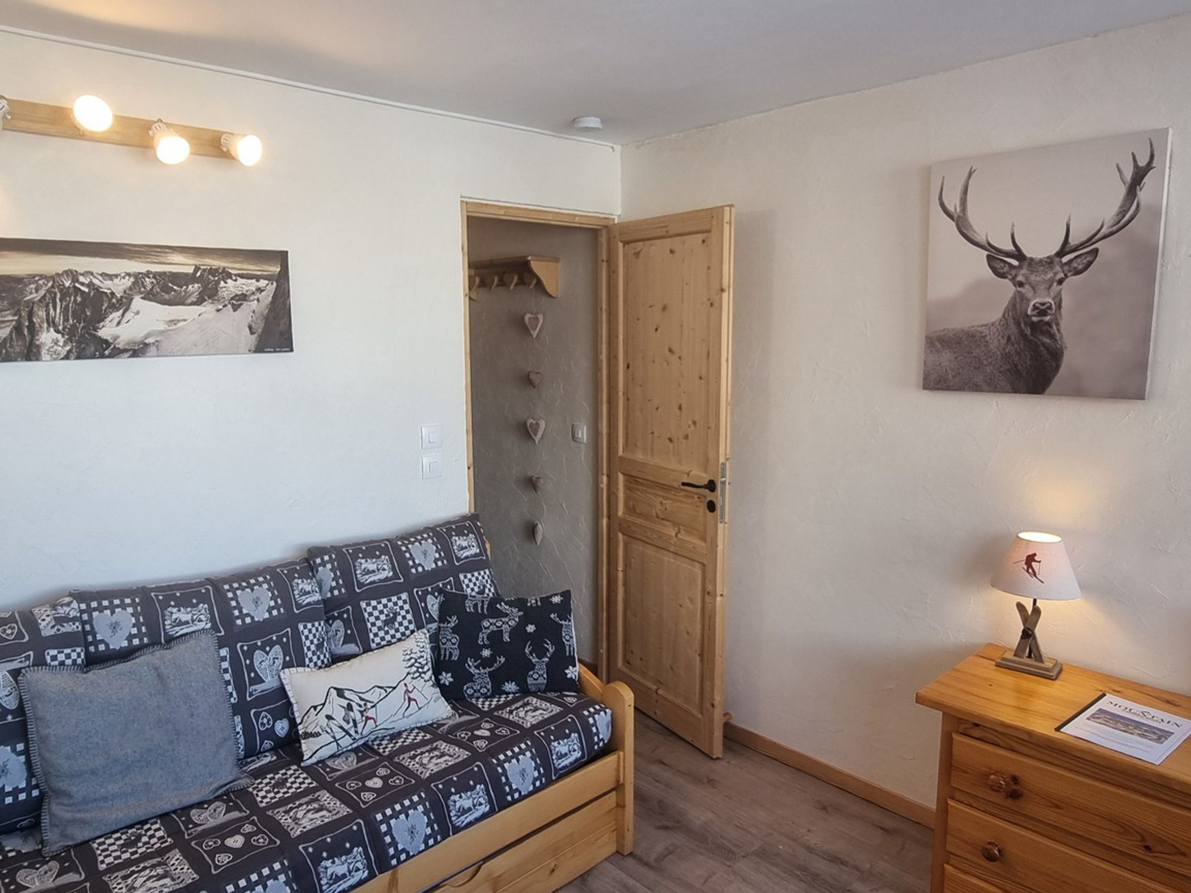 Ein gemütliches Zimmer mit einer gepolsterten Couch, einer Holztür, einer Wandlampe und einem Bild von einem Reh im Bilderrahmen.