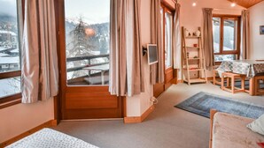 Une pièce chaleureuse avec un plafond en bois, une grande fenêtre avec des rideaux, un lit, un bureau et une vue sur un paysage enneigé.