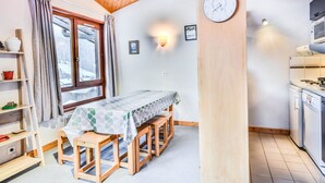 Une pièce chaleureuse avec un plafond en bois, une table à manger avec des bancs, un coin cuisine équipé d’appareils électroménagers et une fenêtre donnant sur un paysage enneigé.