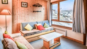 Une pièce chaleureuse avec un mur en bois, un canapé agrémenté de coussins colorés, une petite table et une fenêtre donnant sur un paysage enneigé.