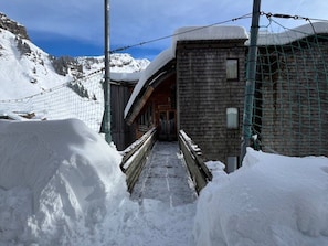 Uma trilha coberta de neve que leva a um edifício de madeira, com uma cerca de metal e montes de neve em ambos os lados.