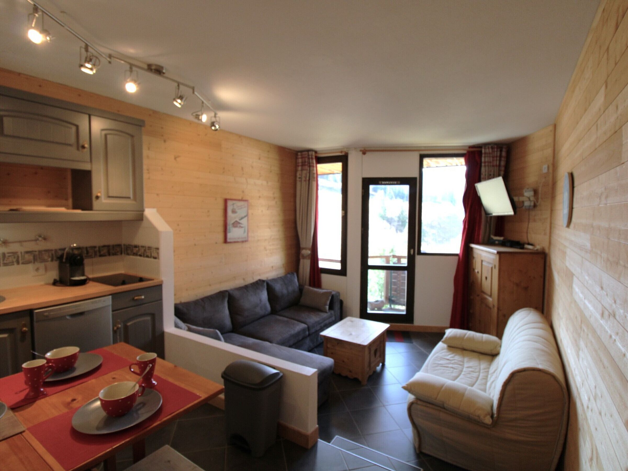 A compact living space with a kitchenette, a sofa, a dining table with red placemats, and a television on a wooden cabinet.