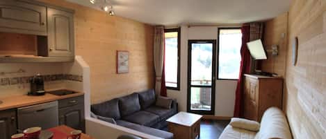 A compact living space with a kitchenette, a sofa, a dining table with red placemats, and a television on a wooden cabinet.