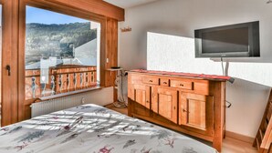 Een slaapkamer met een groot raam, houten meubels, een bed met bloemenbeddengoed en een aan de muur bevestigde televisie.