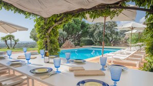 Ein Esstisch für sechs Personen, gedeckt mit blauem Glas, Tellern und Besteck, mit Blick auf einen Pool und eine Lounge-Ecke.