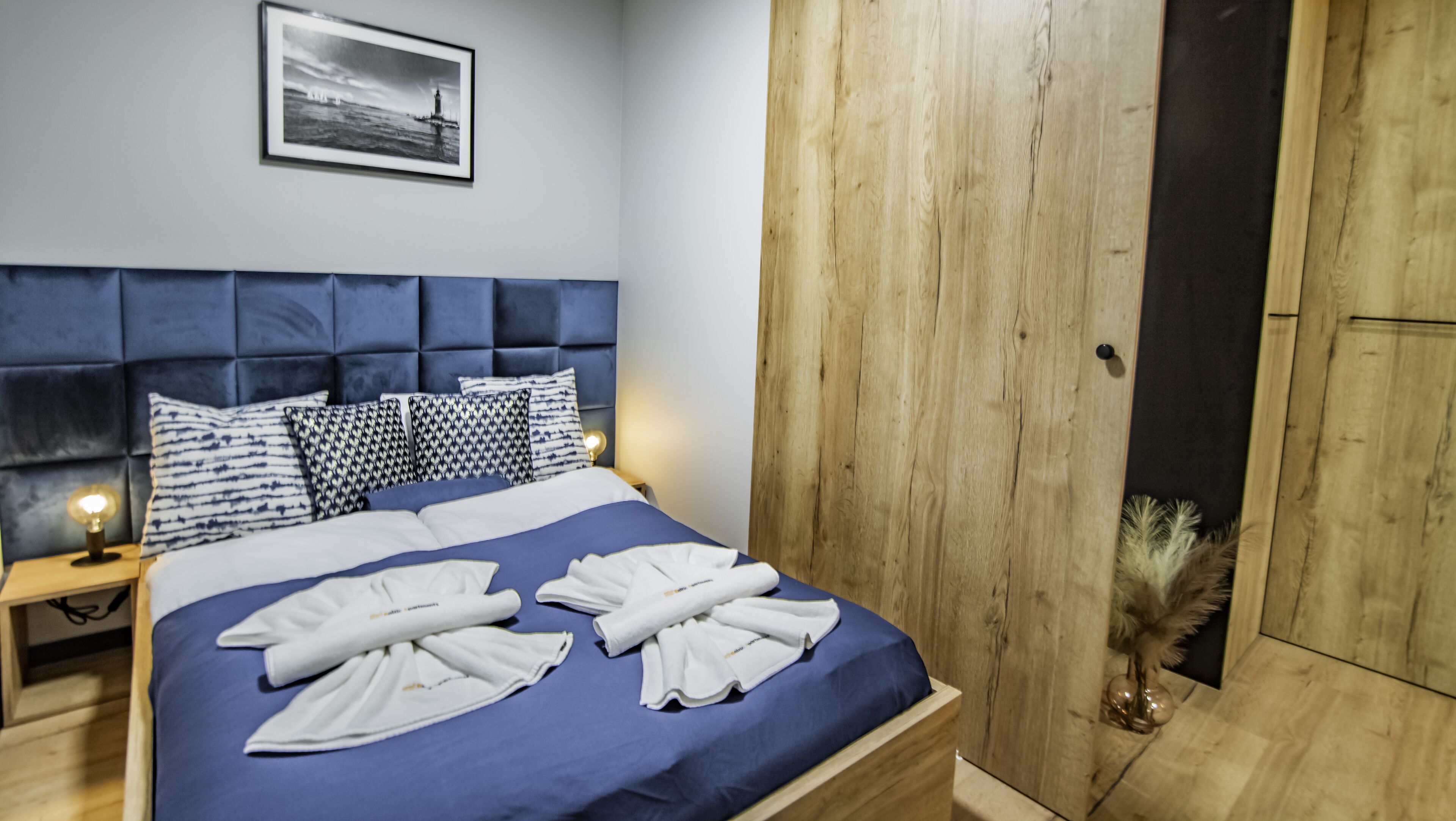 A neatly made bed with blue bedding and white towels, a wooden headboard, and a framed picture on the wall.