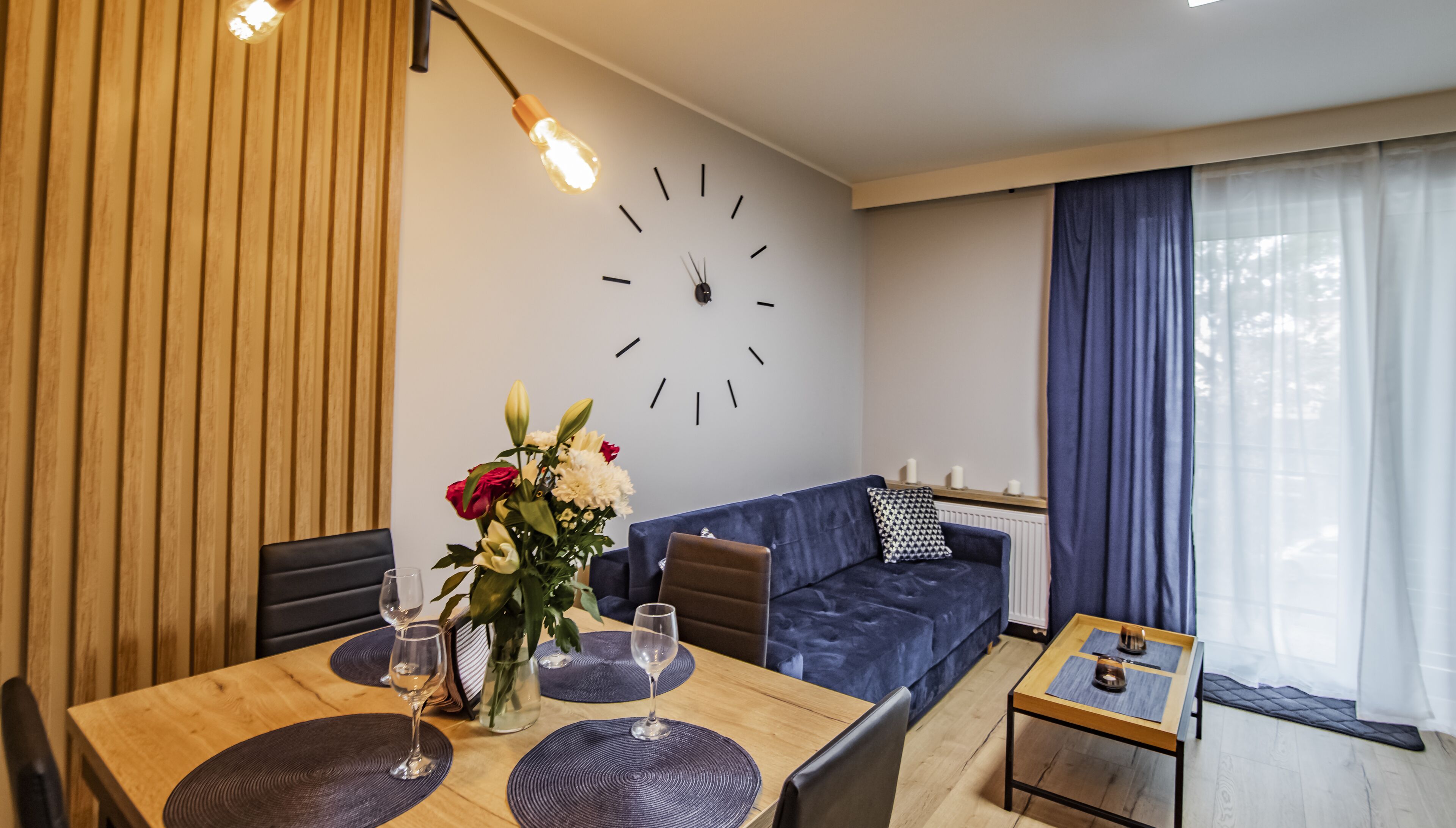 A modern living room with a dining area, featuring a wooden panel wall, a blue sofa, a round clock, and a window with curtains.