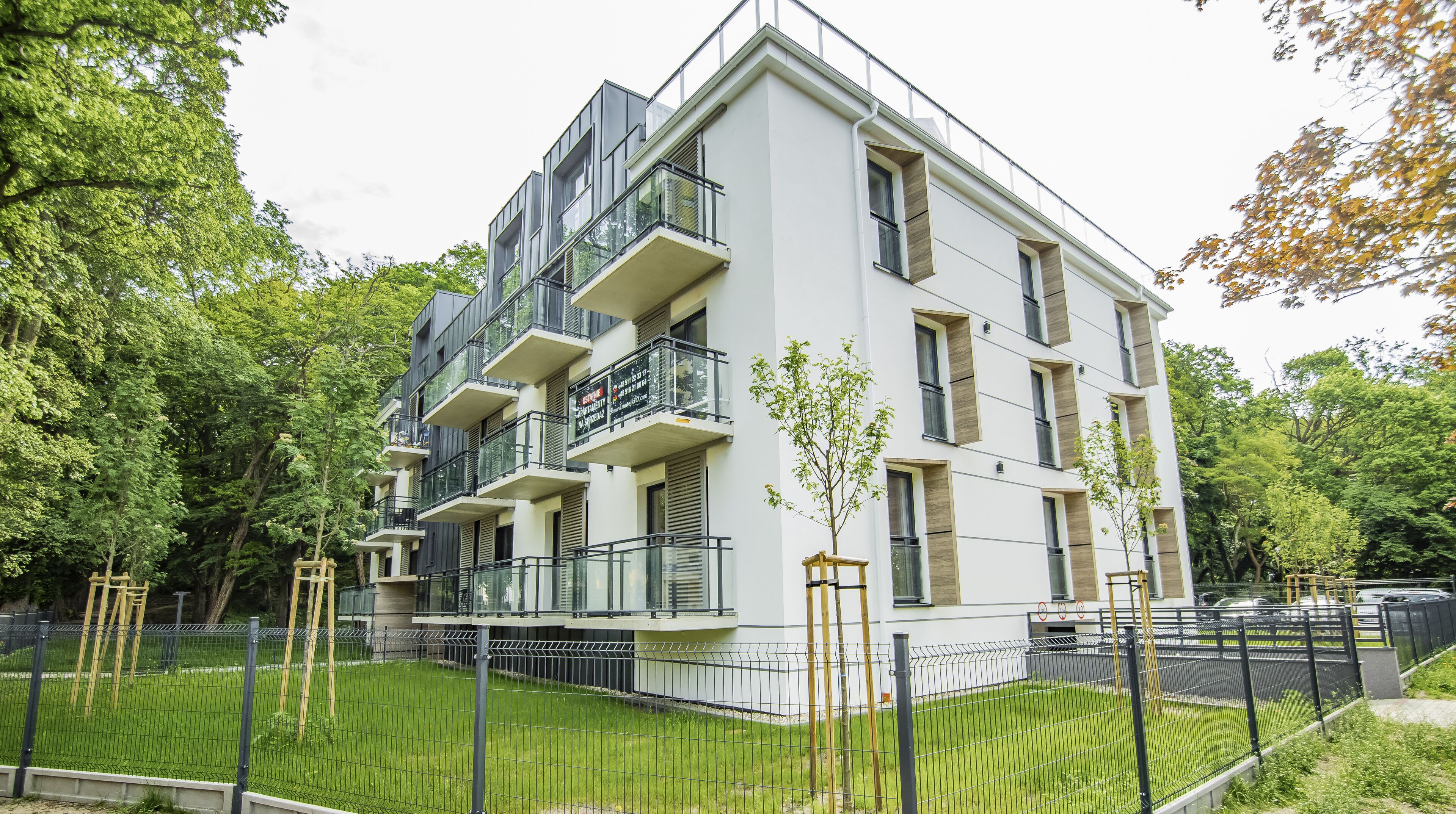 A modern multi-story residential building with balconies, a fenced courtyard, and surrounding greenery.