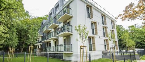 A modern multi-story residential building with balconies, a fenced courtyard, and surrounding greenery.