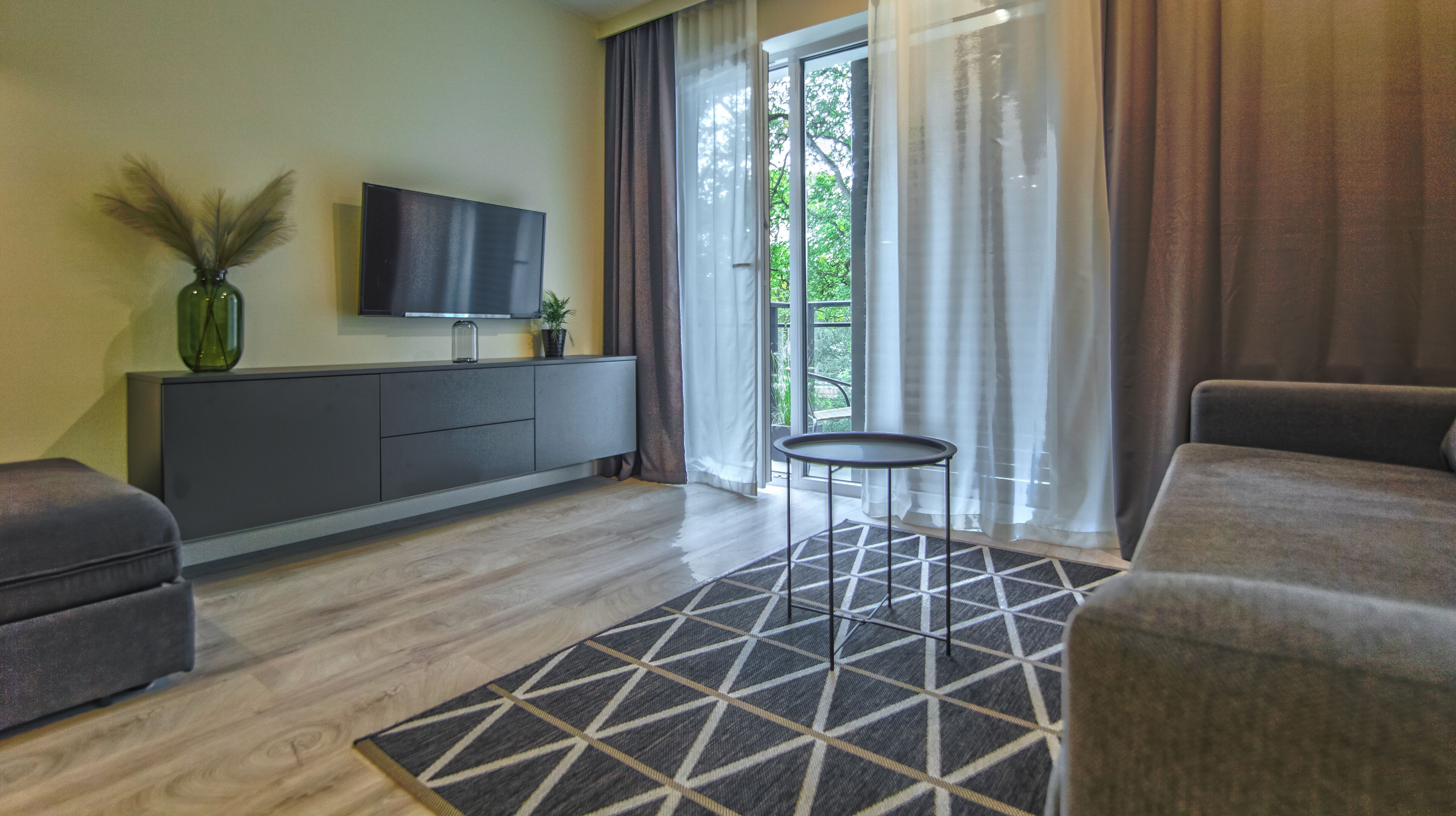 Un salon moderne avec un canapé gris, un téléviseur à écran plat posé sur un meuble, une petite table ronde et un tapis à motifs.