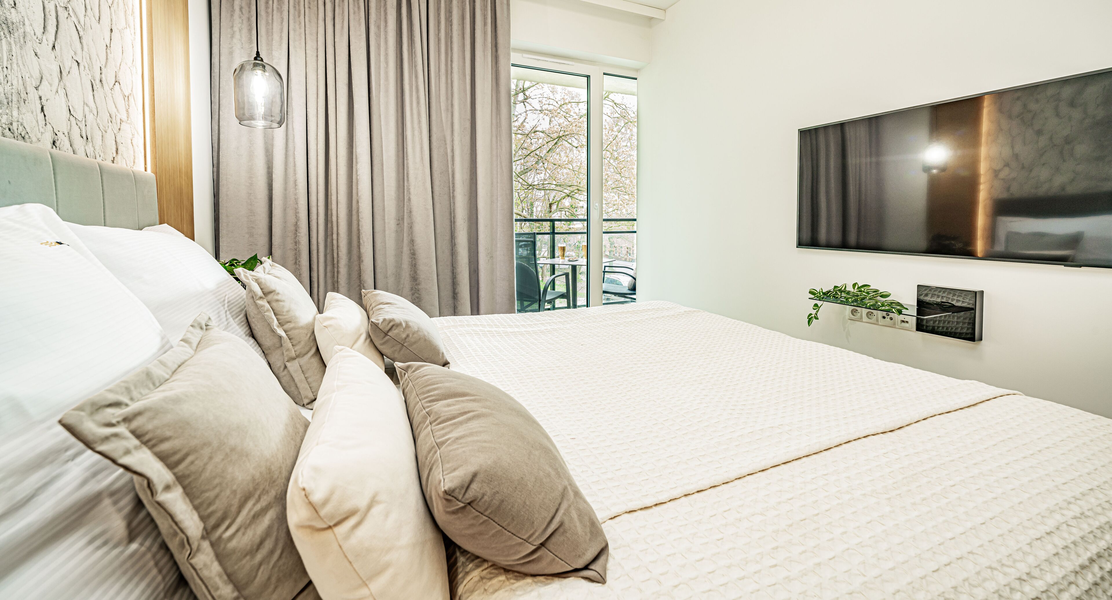 Een netjes opgemaakt bed met beige beddengoed, een modern hoofdbord, een wandtv en een raam met gordijnen.