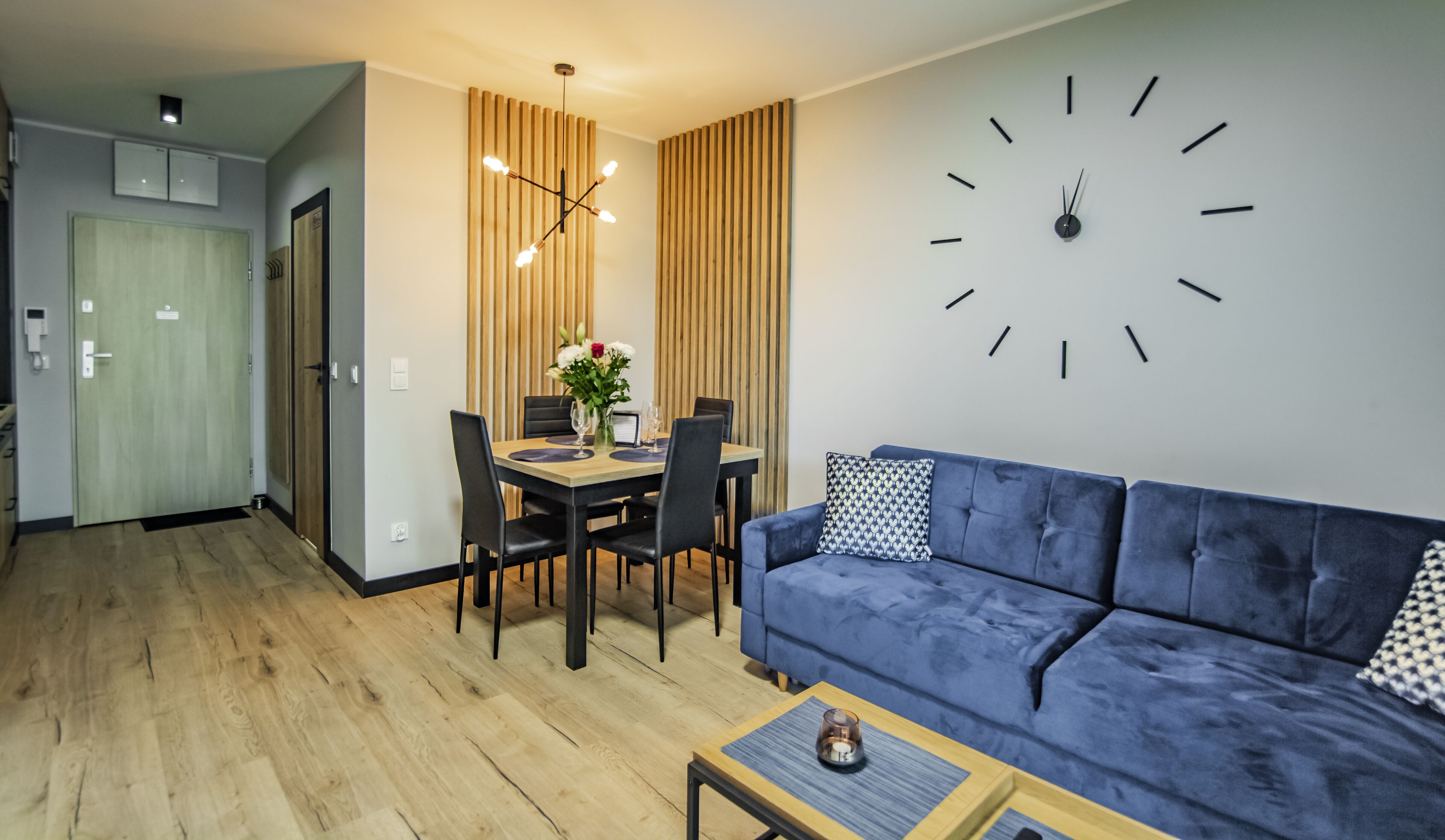 A modern living room with a blue sofa, a dining table with chairs, a wall clock, and a wooden floor.