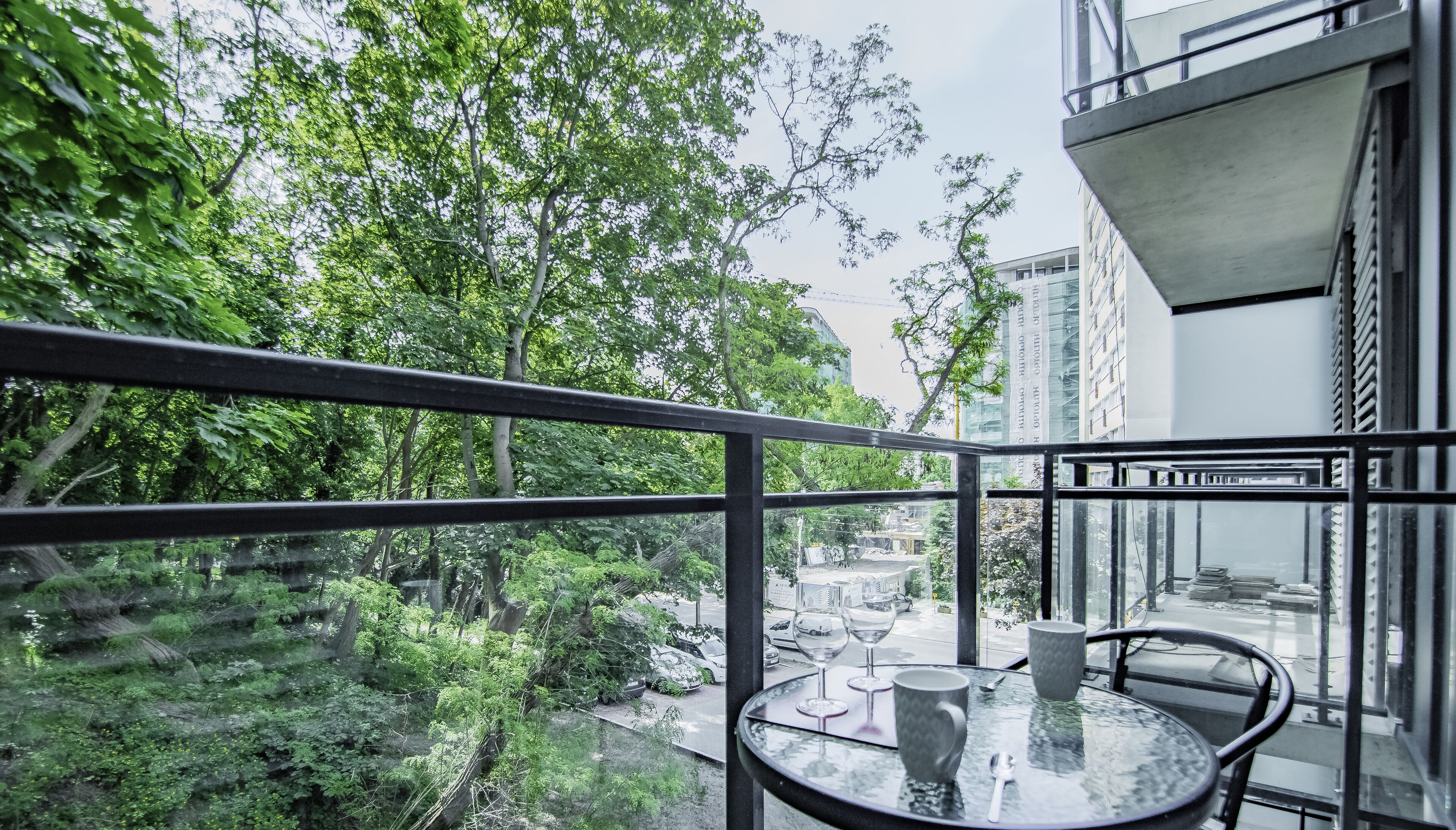 Un balcón con una mesa de vidrio, dos sillas y vistas a un parque verde y edificios.