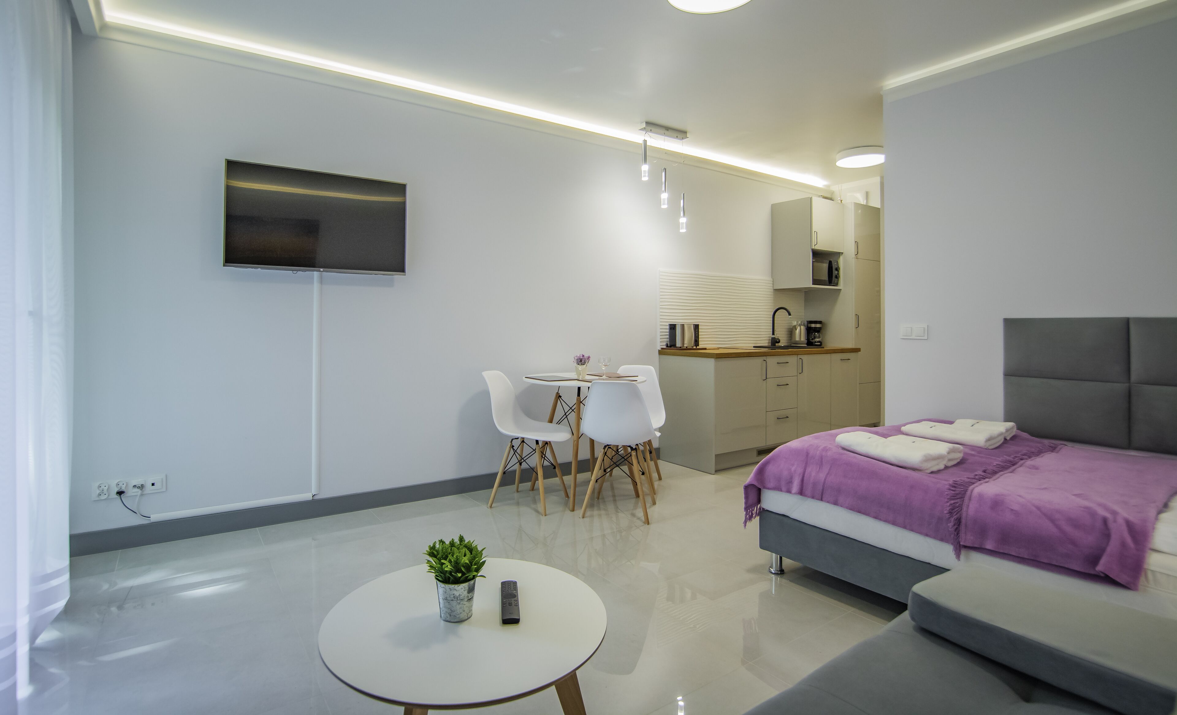 A modern hotel room with a bed, a small dining table with chairs, a kitchenette, and a TV mounted on the wall.