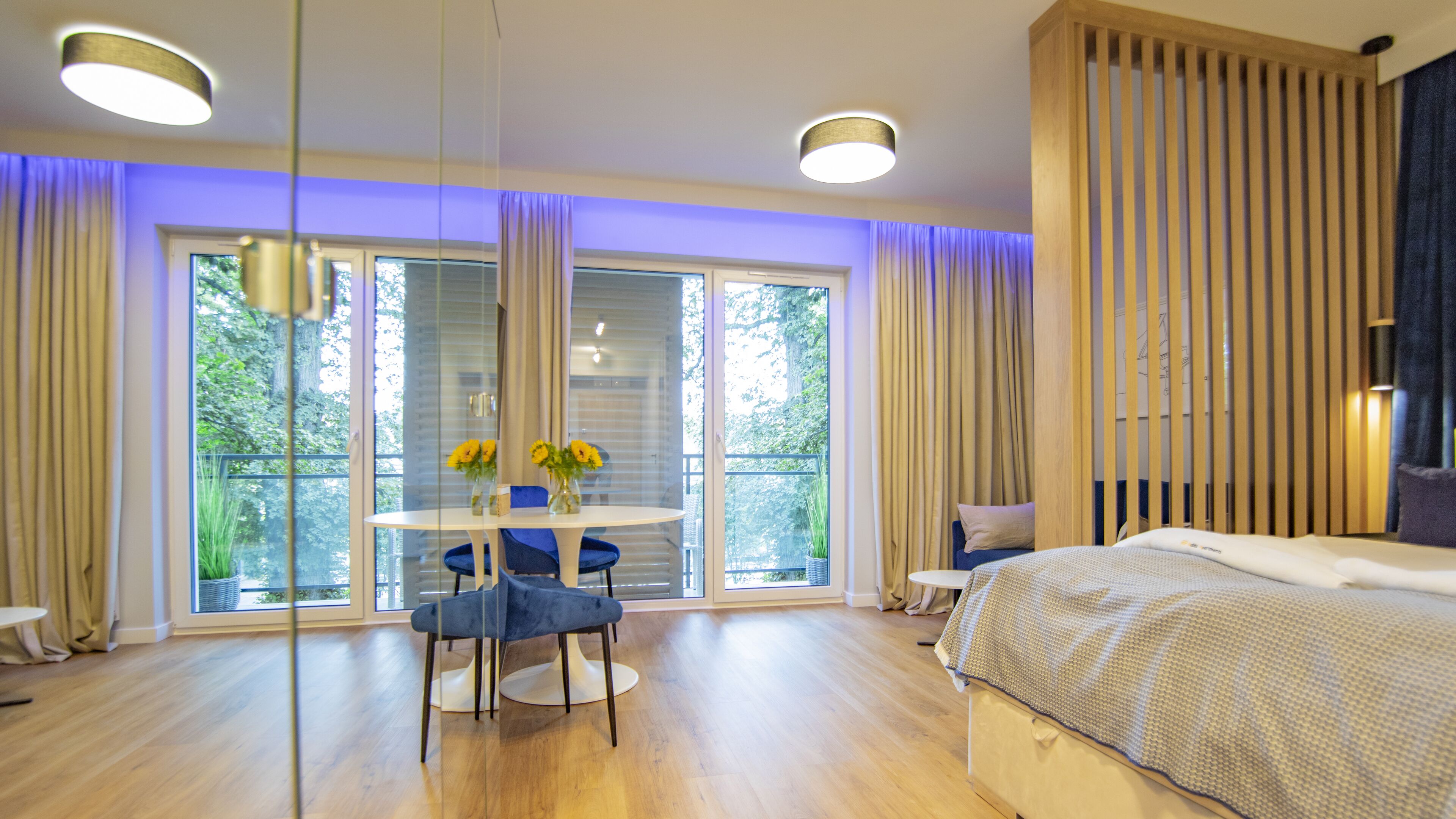 A modern bedroom with a large bed, a round dining table with chairs, and a balcony with curtains.