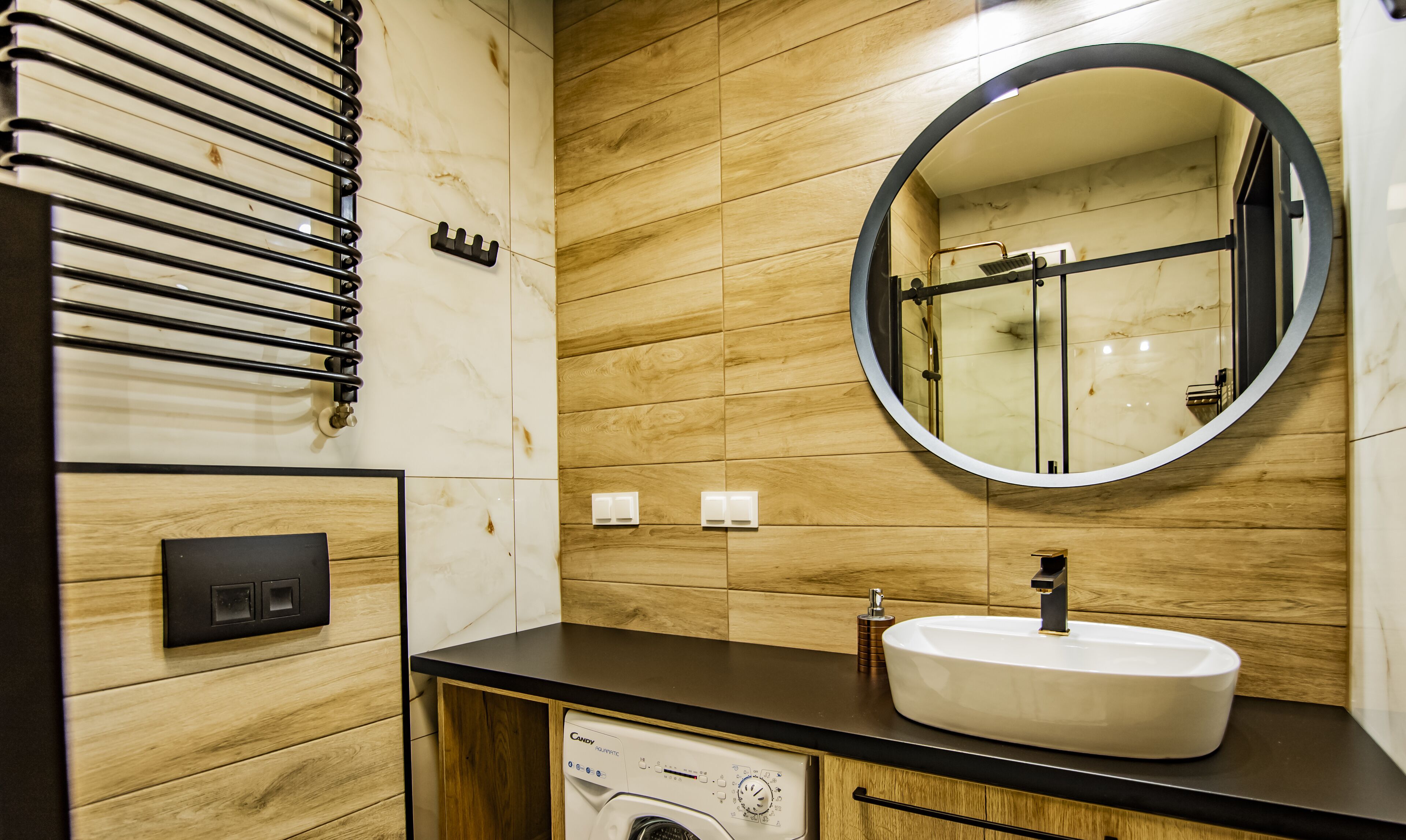 A modern bathroom with a marble wall, a round mirror, a sink, and a built-in washing machine.