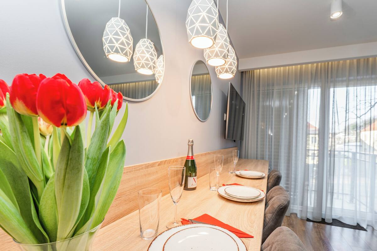 Un comedor moderno con una mesa de madera, servilletas rojas y una botella de champán. Hay tres lámparas colgantes sobre la mesa y un espejo grande en la pared.