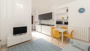 Un appartement moderne doté d’un téléviseur à écran plat posé sur un meuble blanc, d’un coin repas avec des chaises jaunes, d’une cuisine avec des meubles blancs et d’une horloge bleue accrochée au mur.