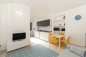 A modern apartment with a flat-screen TV on a white cabinet, a dining area with yellow chairs, a kitchen with white cabinets, and a blue clock on the wall.