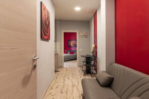 Een gang die leidt naar een slaapkamer met een rode accentmuur, een grijze bank en een zithoek met een tafel en stoelen.