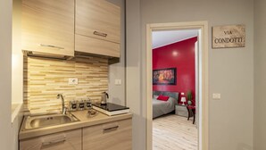Cozinha moderna com bancada de madeira, pia de aço inoxidável e vista para um quarto com paredes vermelhas e uma cama.