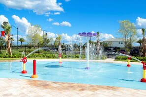 Piscina con varias fuentes de agua, incluyendo fuentes y chorros, rodeada por un patio de baldosas y una valla.