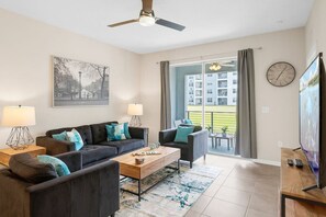 A modern living room with a sofa, armchairs, a coffee table, and a TV. There is a balcony with a view of buildings and a clock on the wall.