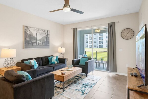 A modern living room with a sofa, armchairs, a coffee table, and a TV. There is a balcony with a view of buildings and a clock on the wall.