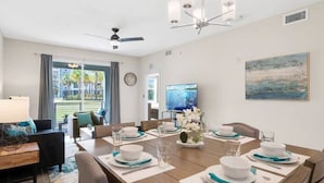 Un comedor con una gran mesa dispuesta para seis personas, un televisor en la pared y vistas al exterior a través de las ventanas.