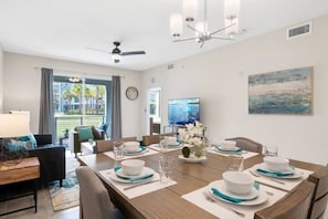 A dining area with a large table set for six, a television on the wall, and a view of the outdoors through the windows.