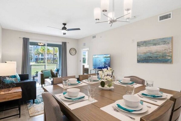 Un coin repas avec une grande table dressée pour six personnes, un téléviseur fixé au mur et une vue sur l’extérieur par les fenêtres.