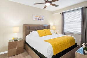 Una habitación de hotel con una cama, dos mesitas de noche, un ventilador de techo, una ventana con cortinas y una obra de arte colgada en la pared.