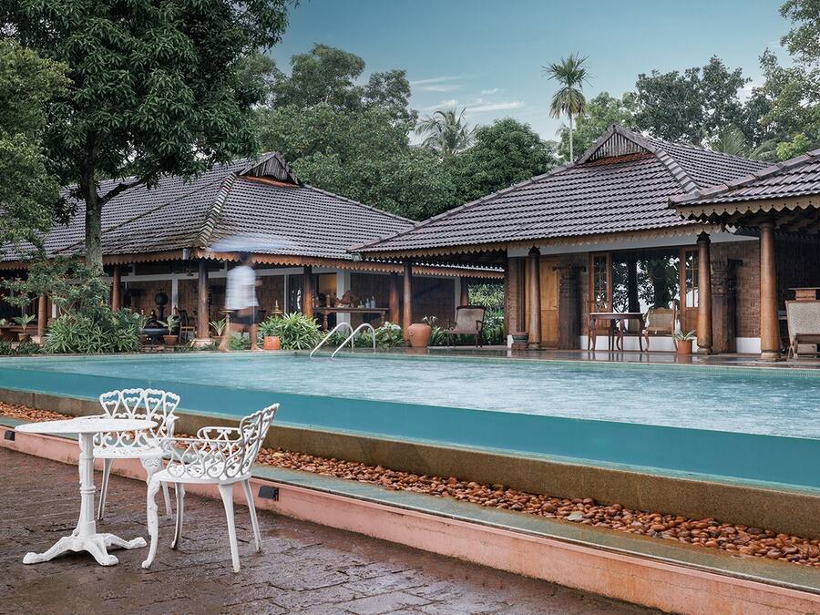 Uma área à beira da piscina com um espaço de estar coberto, piscina e vegetação ao redor.