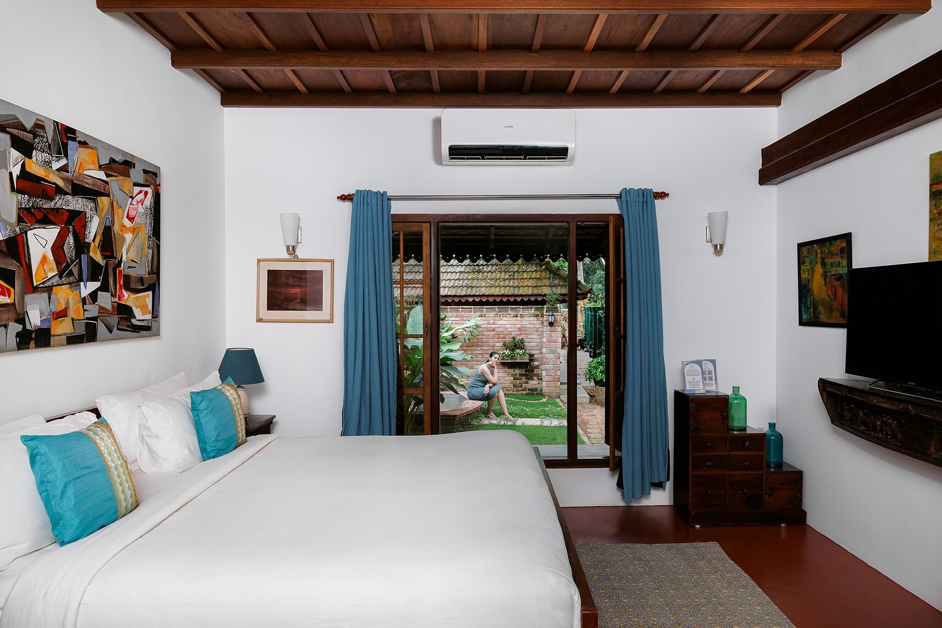 A bedroom with a large bed, a view of a garden, a TV, and artwork on the walls.