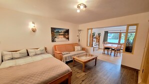 A cozy bedroom with a large bed, a sofa, and a small table. There is a dining area with a table and chairs, and a view of the outdoors through large windows.