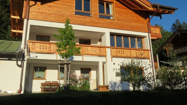 A two-story wooden chalet with balconies, a covered porch, and a small garden area.