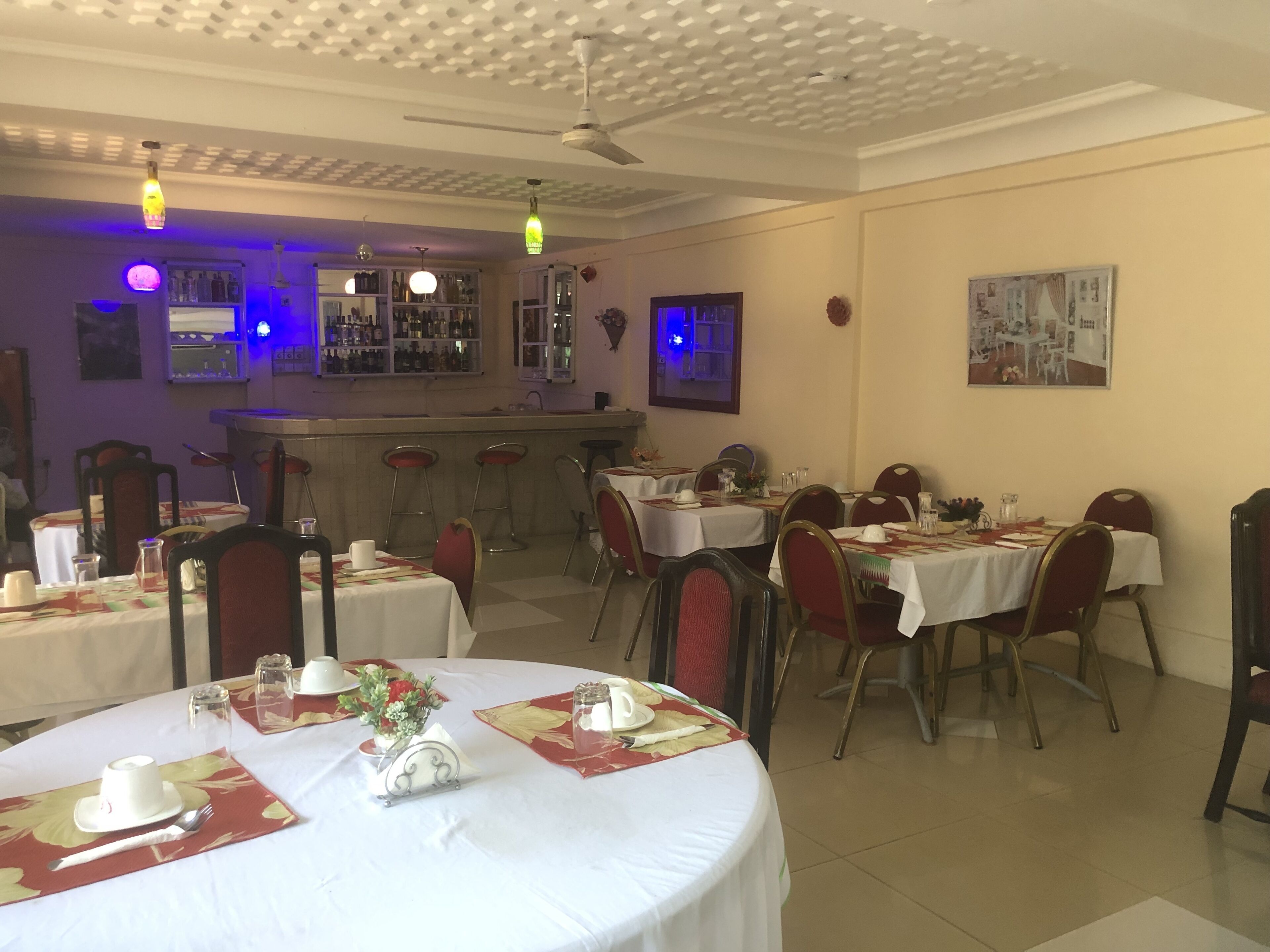 Un restaurante con mesas redondas cubiertas con manteles blancos, salvamanteles rojo y dorado, y sillas rojas. Se ven luces colgantes y un ventilador de techo. En el fondo, se distingue una zona de bar con botellas y vasos.