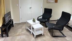 A room with a white door, a small white table with a plant, two black chairs, and a TV on a black stand.