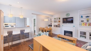 Cozinha moderna com armários brancos, sala de jantar com mesa e cadeiras de madeira, TV montada acima de uma lareira e estante com diversos objetos.