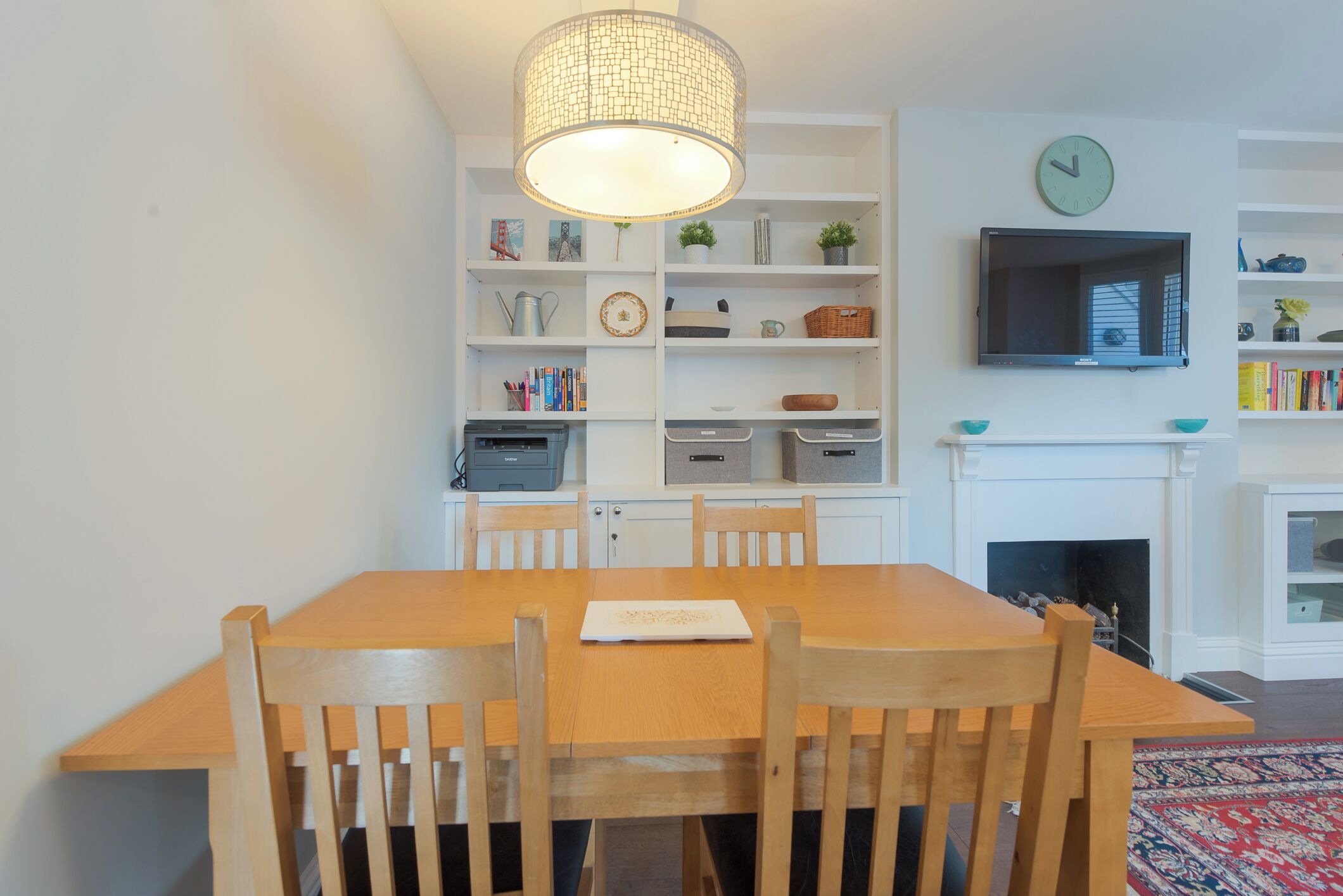 A dining area with a wooden table and chairs, a bookshelf with various items, a television mounted on the wall, and a fireplace.