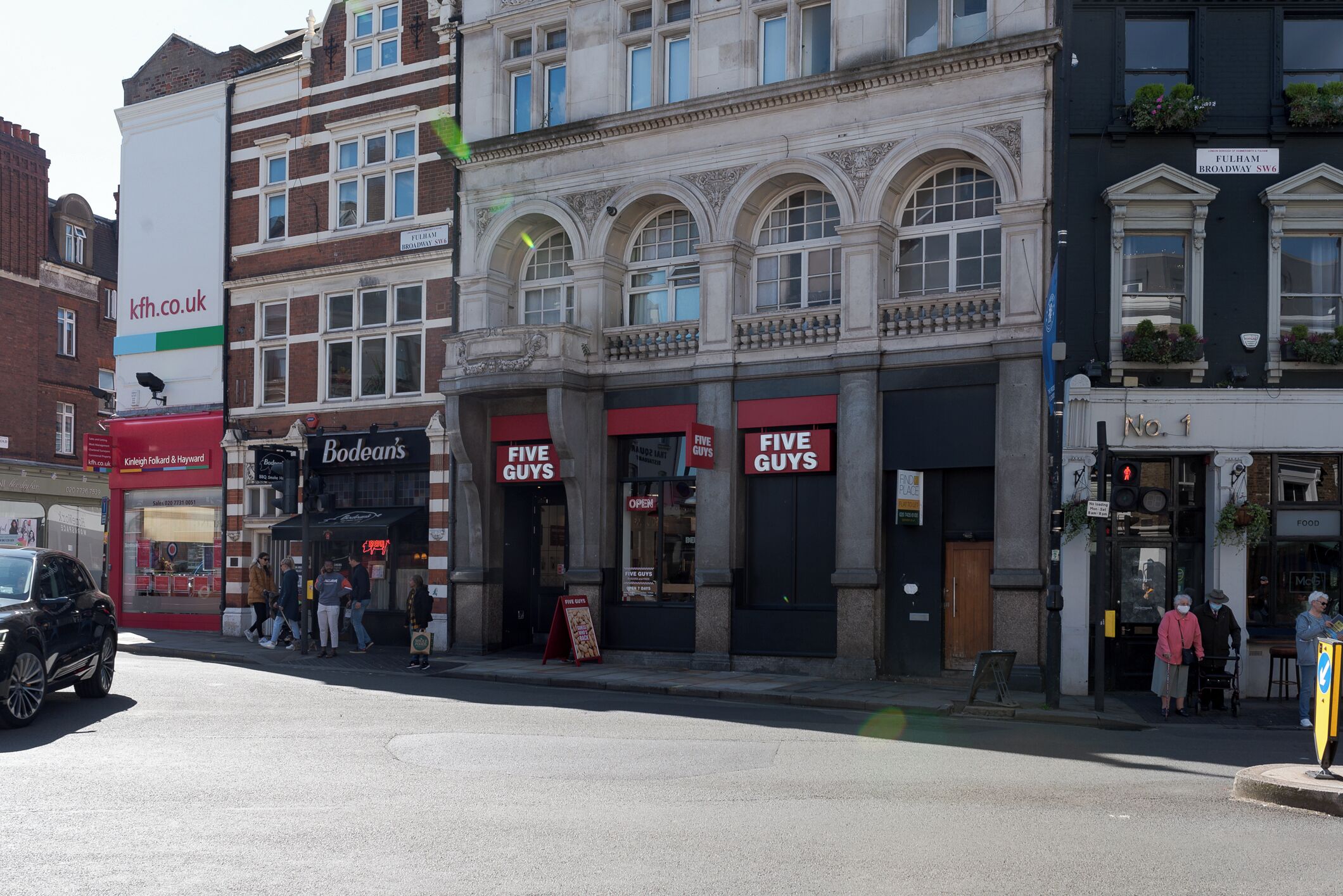 Una vista di strada con un edificio che ospita diverse attività commerciali, tra cui 'Bodeins' e 'Five Guys'.