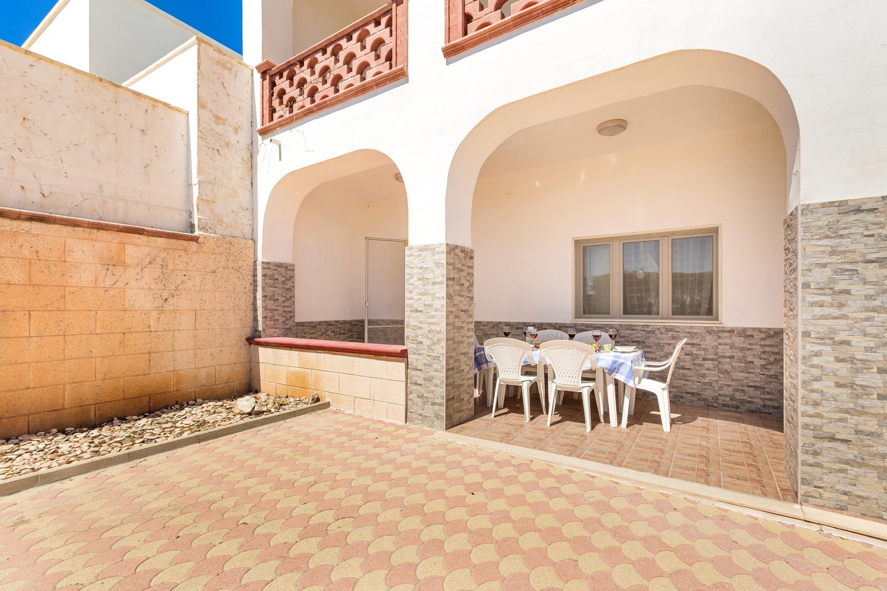 A courtyard with a tiled floor, white chairs, a table with a blue tablecloth, and a window with curtains.