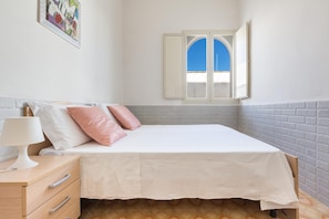 A bedroom with a bed, a nightstand, a lamp, a window with an open shutter, and a wall-mounted artwork.