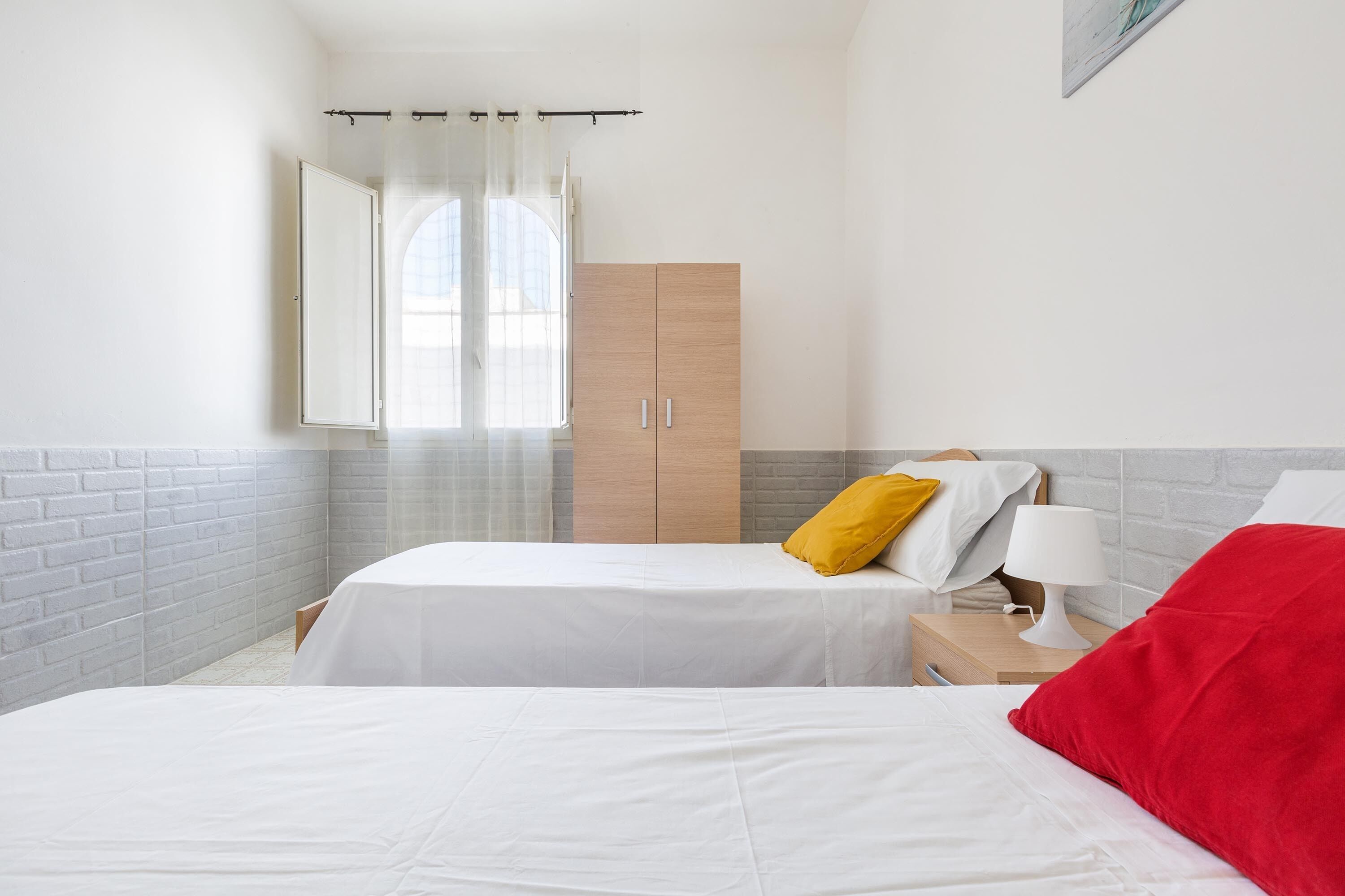 Un dormitorio con dos camas, un armario de madera, una mesita de noche con lámpara y una ventana con cortinas traslúcidas.